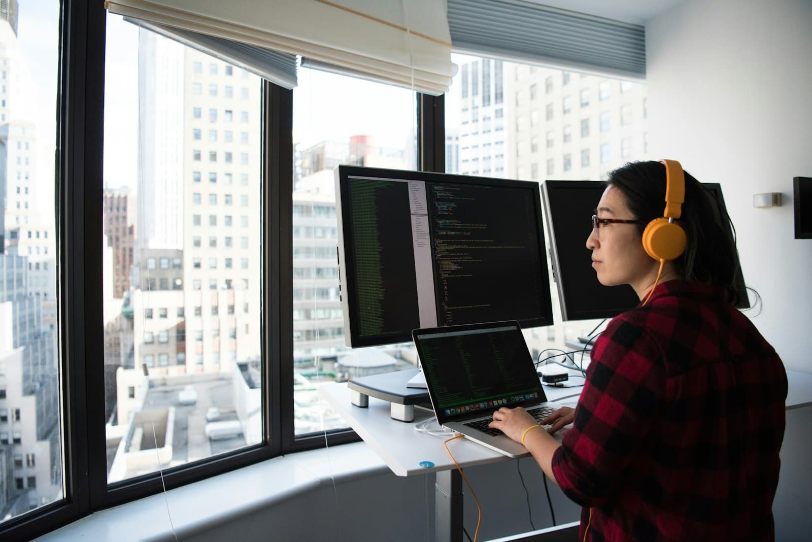 Yui coding at a standing desk by a bright high-rise window, wearing headphones.